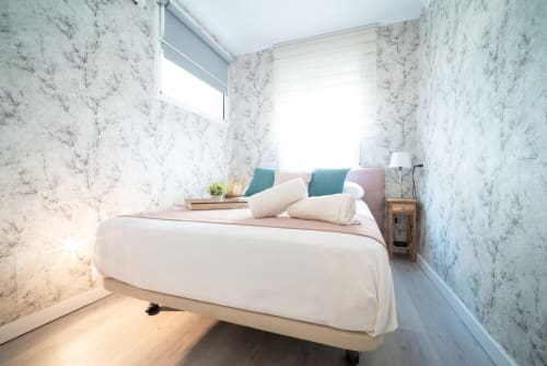 Dormitorio moderno en Luxer Holidays Málaga con paredes de mármol, cama matrimonial, almohadas turquesas, mesita de madera y luz natural