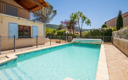 Turquoise swimming pool with wooden deck at villa in Millau, surrounded by gardens and terrace with stone house