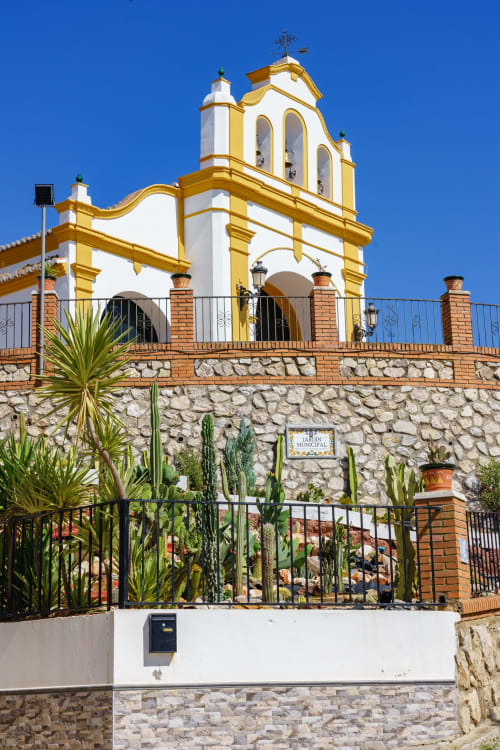 Colonial church with yellow trim and white walls in Colmenar, Spain, featuring a bell tower and cacti garden below