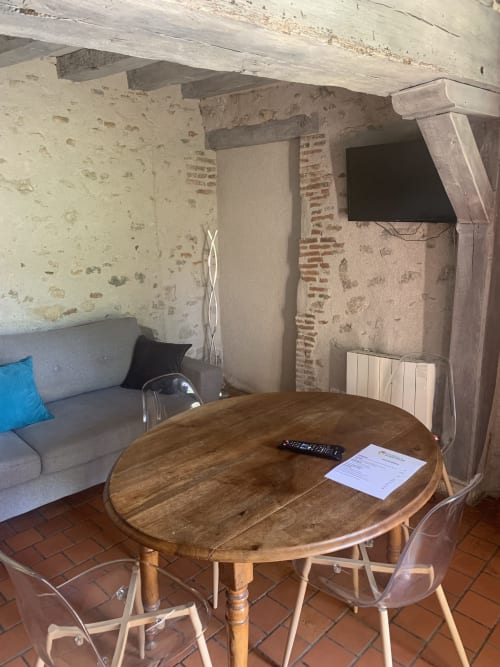 Studio cosy à l'Hôtel De La Bonnheure avec table ronde en bois, canapé gris, TV et murs de pierre rustique à Bracieux