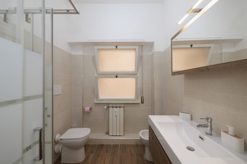 Bagno moderno nell'appartamento ViOletta a Lido di Ostia con lavabo bianco, wc, doccia, finestra e pavimentazione in legno