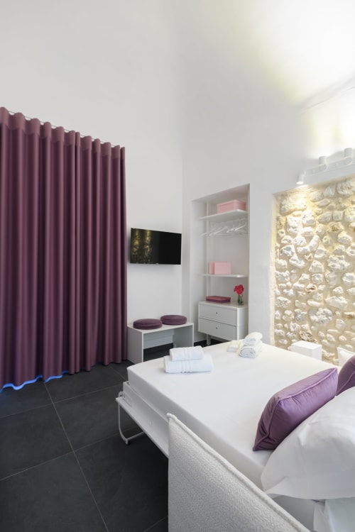 Camera da letto dell'appartamento Peonia all'hotel Almatros di Sulmona con tende viola, letto bianco e parete in pietra