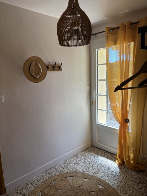 Entrée de la chambre Mimosa au Mas des Vignes avec luminaire suspendu tressé, chapeau de paille, rideaux dorés et sol en terre cuite