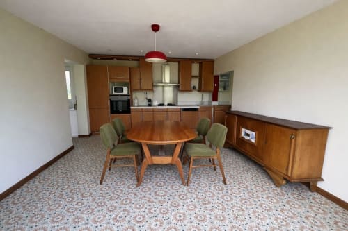Espace salle à manger et cuisine du Petit Verger avec table ronde en bois, chaises vertes, mobilier en bois et luminaire suspendu rouge