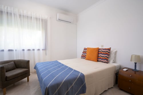 Bright bedroom at RB Algarve hotel in Vilamoura with bed, colorful pillows, blue blanket, sofa, and air conditioning unit