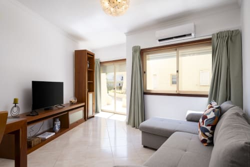 Bright living room in CRIS apartment at RB Algarve Vilamoura with gray sofa, wooden TV stand, glass doors to pool view, and