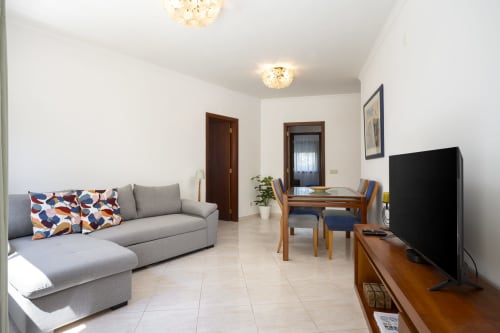 Modern apartment living area at RB Algarve Vilamoura with gray sofa, dining table, TV, and bright natural lighting