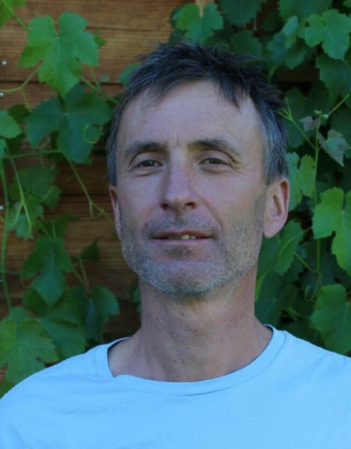 Homme d'âge moyen en chemise bleu clair posant devant un mur en bois avec du lierre grimpant à L'encabanée, gîte et chambre d'hôtes à