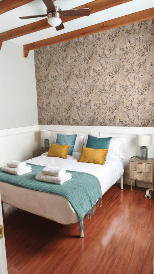 Dormitorio Medieval Mirador con vigas de madera, papel tapiz floral, almohadas teal y amarillas, ropa de cama blanca y pisos de madera