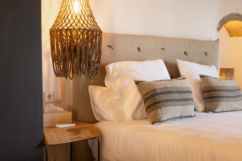 Dormitorio moderno en Mas Escarrà con cabecero tapizado en beige, almohadas blancas, cojines a rayas y lámpara de mimbre