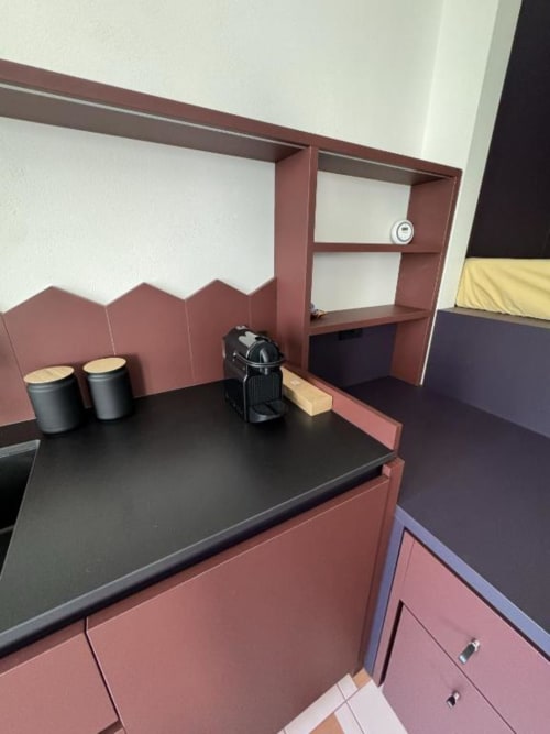 Angolo cottura dello studio Muratori Apartments Milano con pensili bordeaux, piano nero, scaffalature e contenitori per caffè