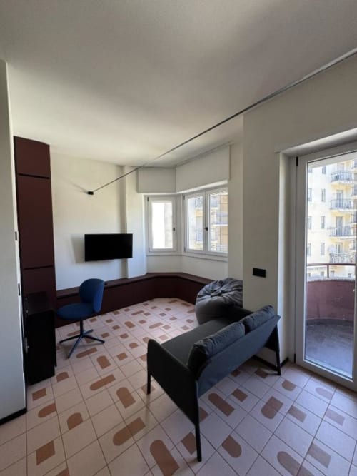 Moderno studio con balcone presso Muratori Apartments Milano, divano nero, scrivania e pavimento in piastrelle decorative.