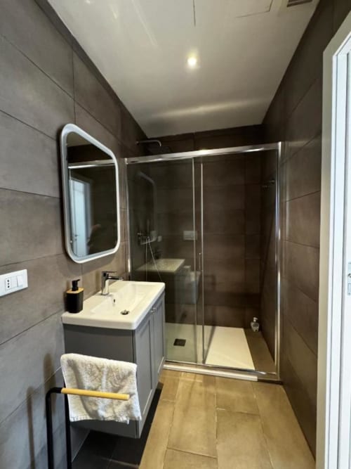 Bagno moderno nell'appartamento milanese con piastrelle scure, lavabo bianco, specchio a LED e box doccia in vetro