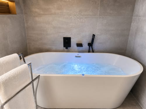 Moderna bañera exenta blanca con agua iluminada en azul y accesorios negros en el hotel Holibai de Baiona, España