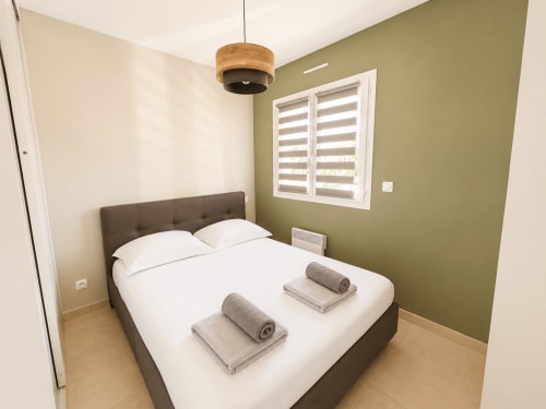 Chambre côtière douillette à Casa Di Bravone avec lit gris, draps blancs, serviettes roulées, mur accentué olive, luminaire suspendu en bois