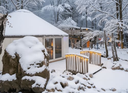 Snow-covered glamping tent with lit windows at Les Murmures du Bois, surrounded by frost-laden trees in winter garden