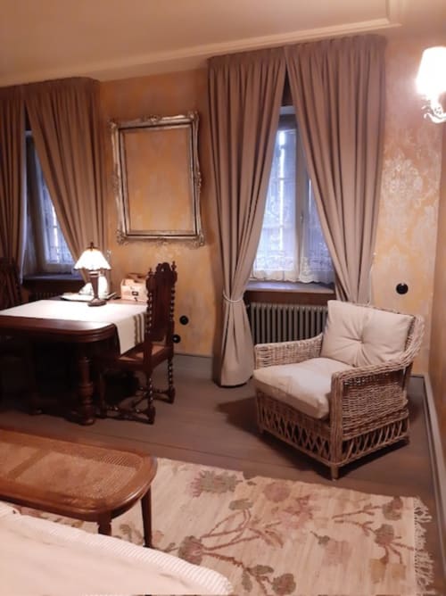 Chambre élégante à la maison oubliée de Goetzenbruck avec mobilier classique, fauteuil en rotin et vue sur le jardin