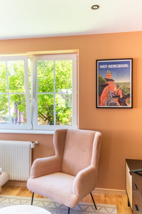 Acogedora habitación de apartamento con paredes color durazno, butaca beige, gran ventana con vistas a árboles verdes y póster de Haut-Kœnigsbourg