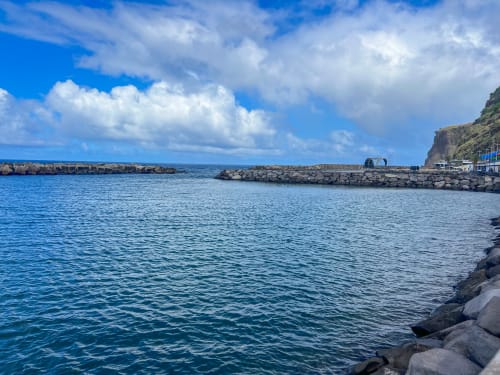 Vista panorâmica do porto costeiro de Funchal com águas calmas e azuis, quebra-mar rochoso, e edifícios à beira dos penhasco sob céu parcialmente nublado