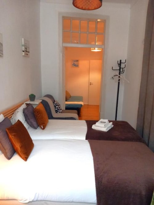 Modern bedroom in Lisbon apartment with white bed, orange accents, seating area, and city views through glass doors