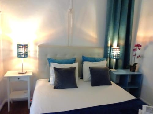Modern bedroom in Lisbon apartment with queen bed, blue headboard, dark pillows, white nightstand with lamp, city view, and