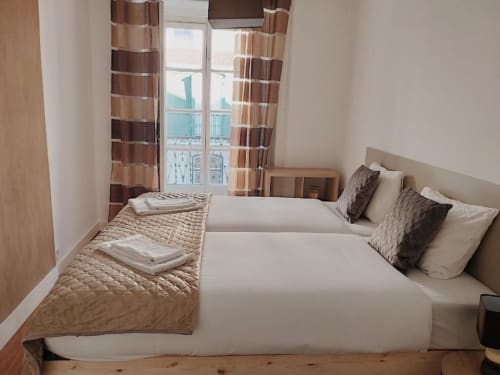Quarto iluminado em apartamento em Lisboa com duas camas, cortinas listradas, assoalho de madeira e vista para a cidade desde a varanda