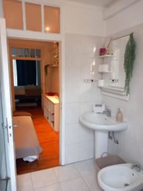 Bright bathroom in Lisbon BnB apartment with white sink, toilet, wall shelving, and orange hallway view