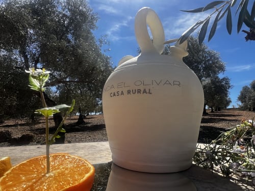 White ceramic jug with 'Finca El Olivar Casa Rural' text surrounded by olive trees and orange slice in rural Spanish garden