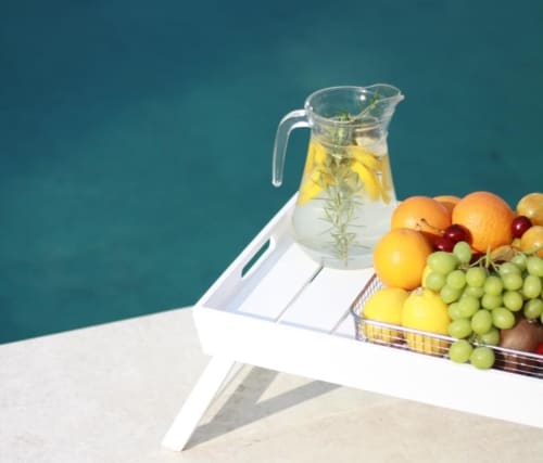 Glass pitcher with infused water and fresh fruit arrangement including grapes and lemons on white table with pool view at