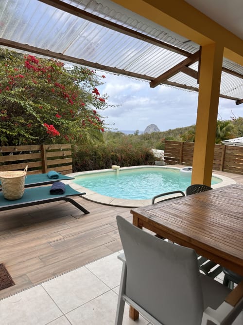 Terrasse de villa tropicale avec piscine turquoise, chaises longues en bois, table à manger et bougainvillées fleuries