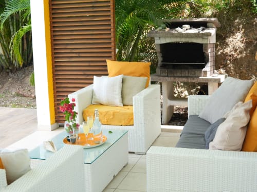 Terrasse de villa tropicale aux Exotic Villas Martinique avec mobilier en osier, coussins jaunes, barbecue en pierre et verdure luxuriante