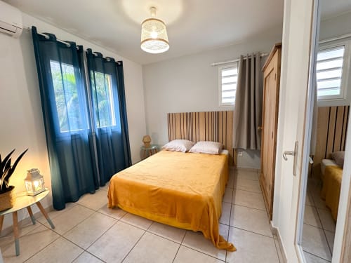 Chambre lumineuse avec couvre-lit jaune, tête de lit en bois, rideaux bleus et vue sur mer aux Exotic Villas Martinique au Diamant