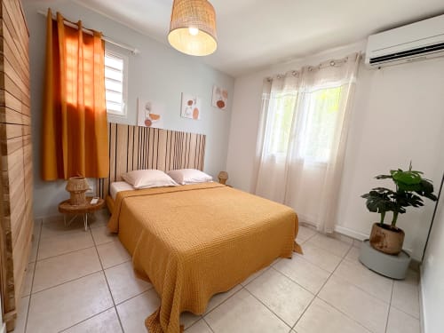 Chambre lumineuse aux Exotic Villas Martinique avec lit jaune, luminaire suspendu en rotin, rideaux orange et plante en pot