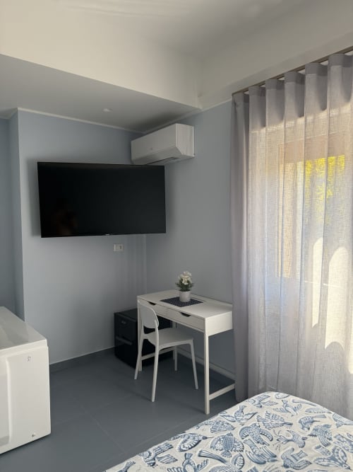 Camera moderna al BellDuomo Suite Messina con TV a parete, scrivania bianca, sedia e biancheria patterned dietro tende trasparenti