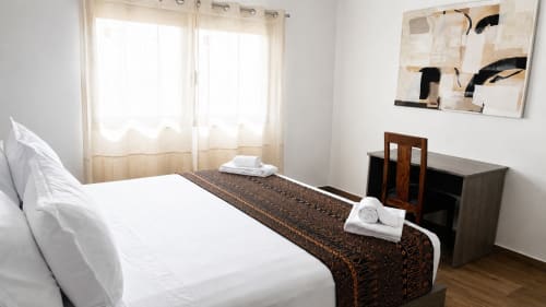 Modern bedroom in Dakar apartment with white bedding, brown runner, wooden desk, abstract art, and city views through sheer
