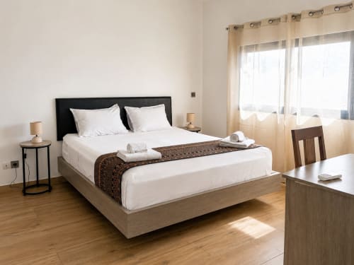 Bright bedroom in Dakar apartment with queen bed, black headboard, white linens, wooden desk, and city view balcony