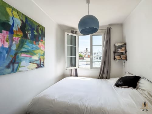 Chambre lumineuse aux Appartements Montmartre Paris avec lit blanc, œuvres d'art colorées, lampe suspendue bleue et vue sur la ville