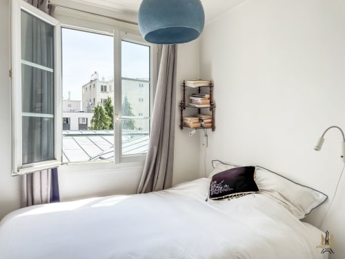 Chambre lumineuse aux Appartements Montmartre Paris avec lit blanc, luminaire bleu suspendu, larges fenêtres avec vue sur les toits de la ville