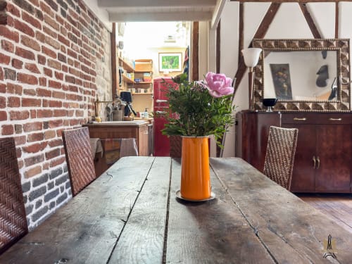 Salle à manger avec table en bois, vase orange avec fleurs roses, mur en brique, et chaises tissées aux Appartements Montmartre Paris