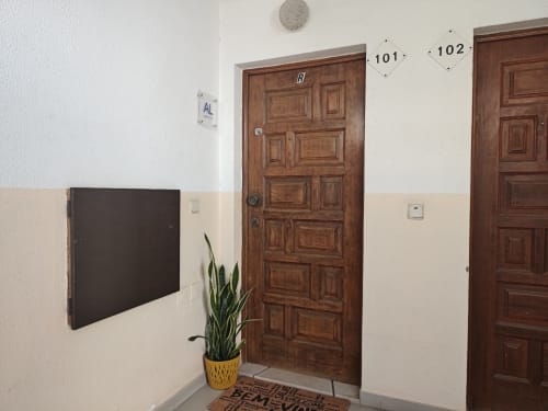 Entrada do corredor da Albufeira Inn com porta de madeira, número do quarto 101, planta em vaso e tapete de boas-vindas