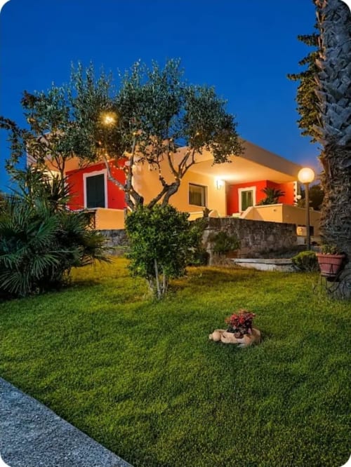 Dimora Pierino villa at dusk with illuminated red and yellow exterior, manicured garden, olive trees, and landscape lighting