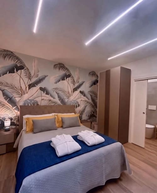 Camera moderna presso Anima Est B&B Napoli con murale di foglie di palma, luci al neon a soffitto, biancheria blu e pavimenti in legno