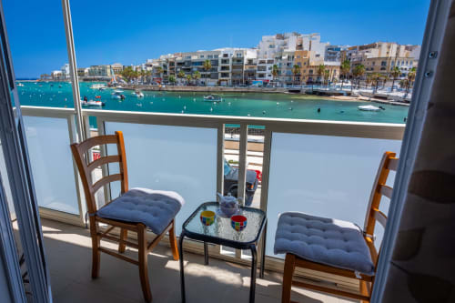 Vista dal balcone della baia turchese con sedie di legno e tavolino che si affaccia sul porto di Marsaskala, edifici colorati sul lungomare