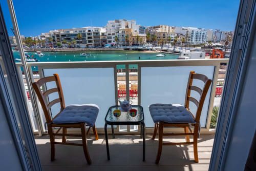 Balcone sul lungomare con sedie in legno che si affacciano sulle acque turchesi del Mediterraneo e gli edifici costieri di Marsaskala, Malta