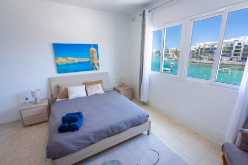 Luminosa camera da letto sul lungomare con letto matrimoniale, finestra con vista sul porto di Marsaskala e opere d'arte del paesaggio costiero.