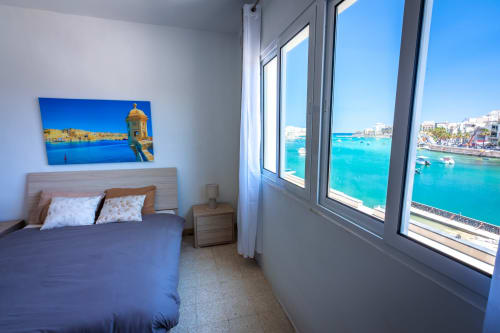 Camera con vista sul mare turchese attraverso grandi finestre, letto blu e opere d'arte costiera incorniciate presso Calm Holiday Homes,