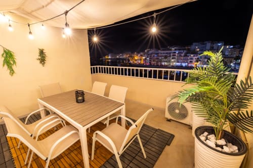Terrazza sul mare della casa di Marsaskala con tavolo da pranzo, sedie, piante e vista notturna del mare e degli edifici costieri