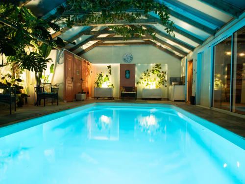 Piscine couverte L'Écrin des Roches avec eau bleue, toit en verre, plantes, terrasse en bois et espace détente