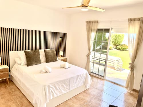 Dormitorio moderno en villa de Nerja con cabecero de rayas, ropa de cama blanca, ventilador de techo y vistas al jardín a través de puertas de cristal