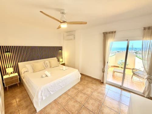 Dormitorio moderno en villa de Nerja con vistas al mar, cama blanca, ventilador de techo de madera, acceso a terraza y paisaje costero visible